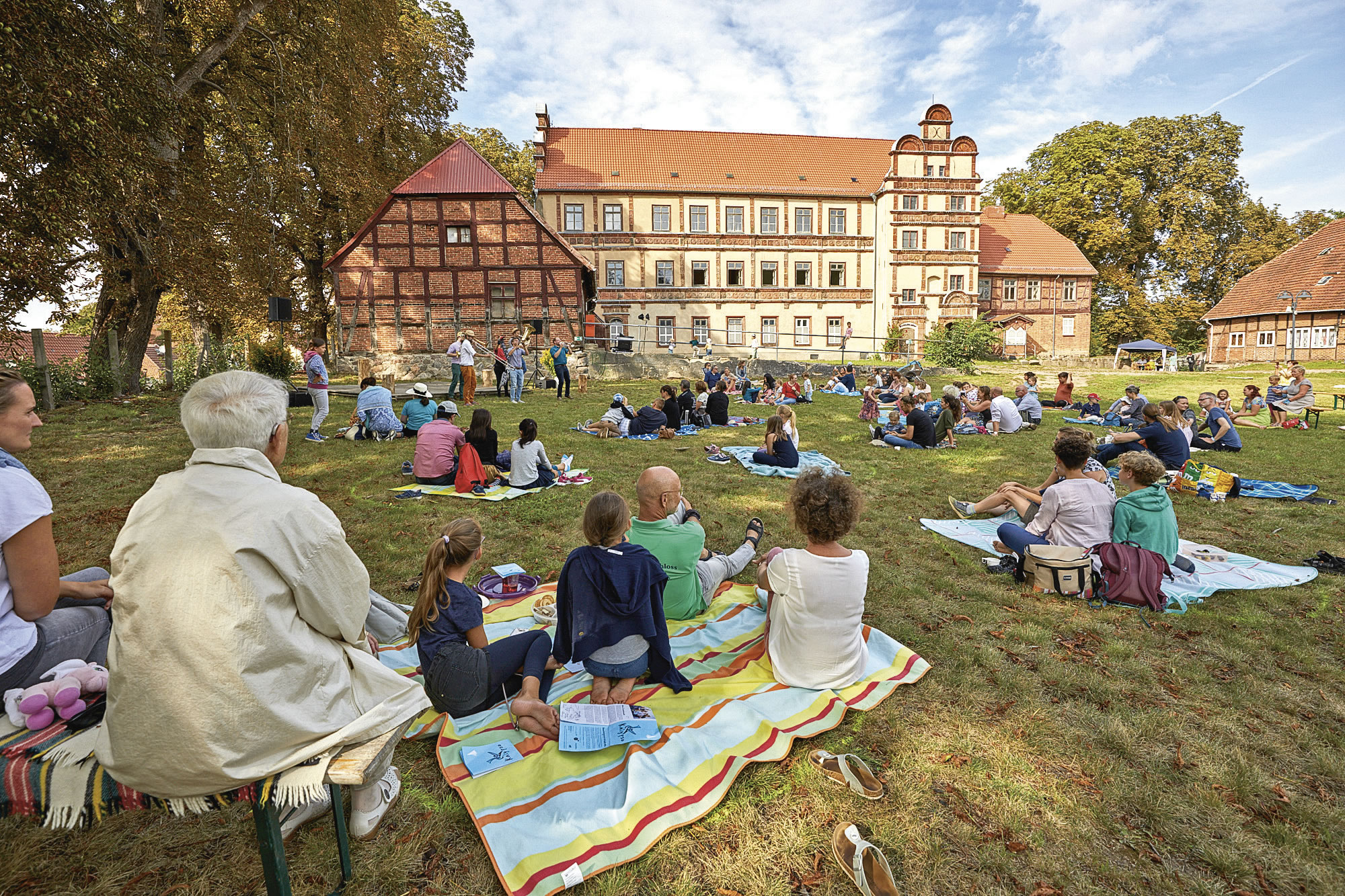 Auf einer Wiese picknicken einige Gäste, im Hintergrund ein Fachwerkschloss. Vor dem Gebäude eine Band.