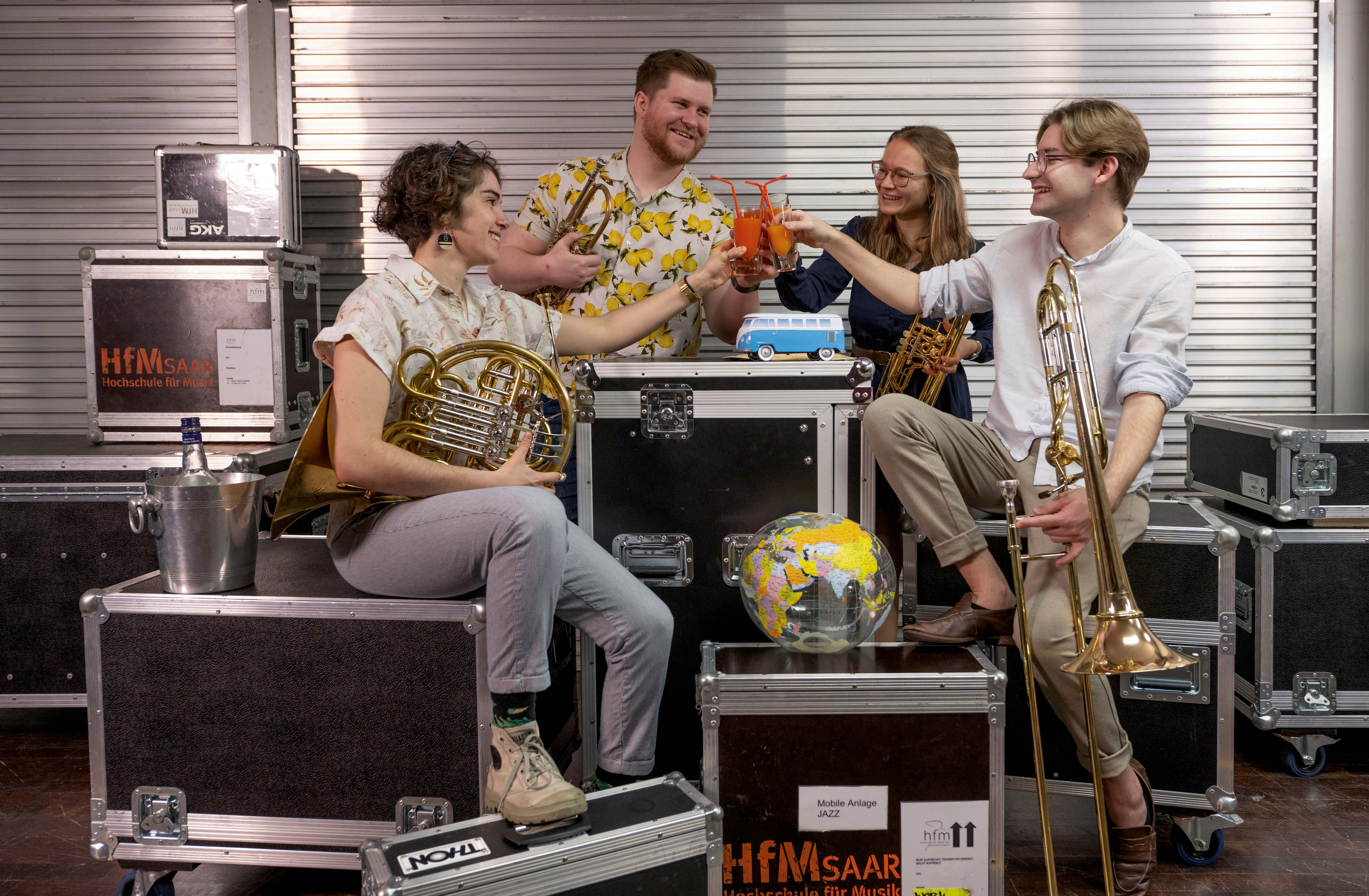 Musikstudierende sitzen auf mehreren Transportkisten, habe ihre Instrumente in der Hand und stoßen mit Cocktails an.