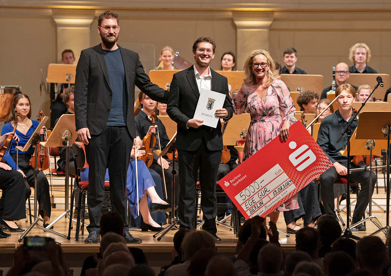 Die genannten Personen im niedrigeren mittleren Alter stehen in Anzügen und Kleid vor einem Orchester. Sarah Wedl-Wilson hält einen großen Scheck in der Hand.