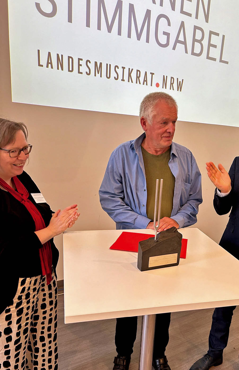 Ein Mann etwas höheren mittleren Alters in einem blauen Hemd mit kurzer weißer Halbglatze steht vor seiner Auszeichnung: Der Goldenen Stimmgabel