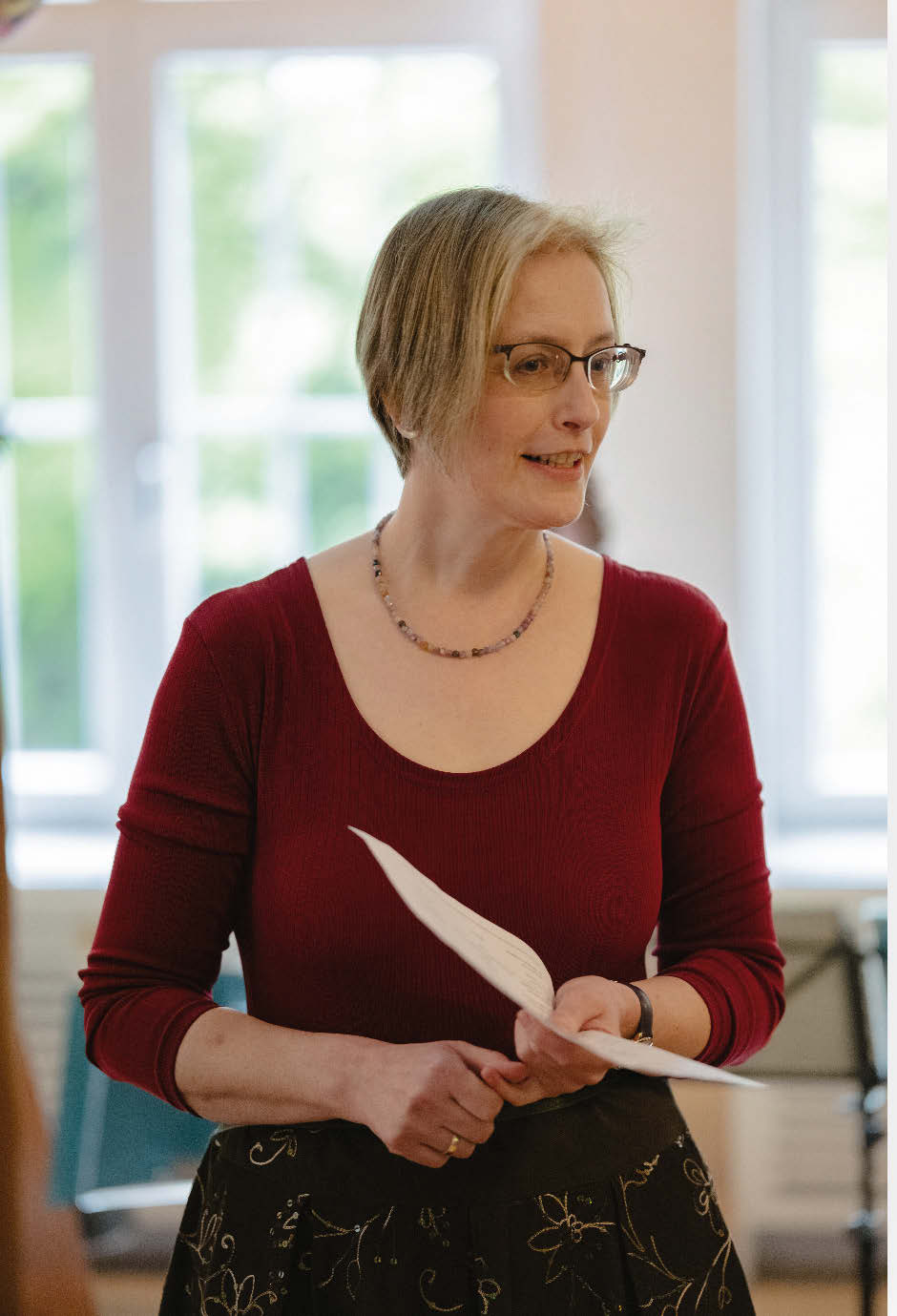 Eine Frau mittleren Alters mit glatten halblangen blonden Haaren und einem roten Oberteil spricht in einem kleineren Probensaal.