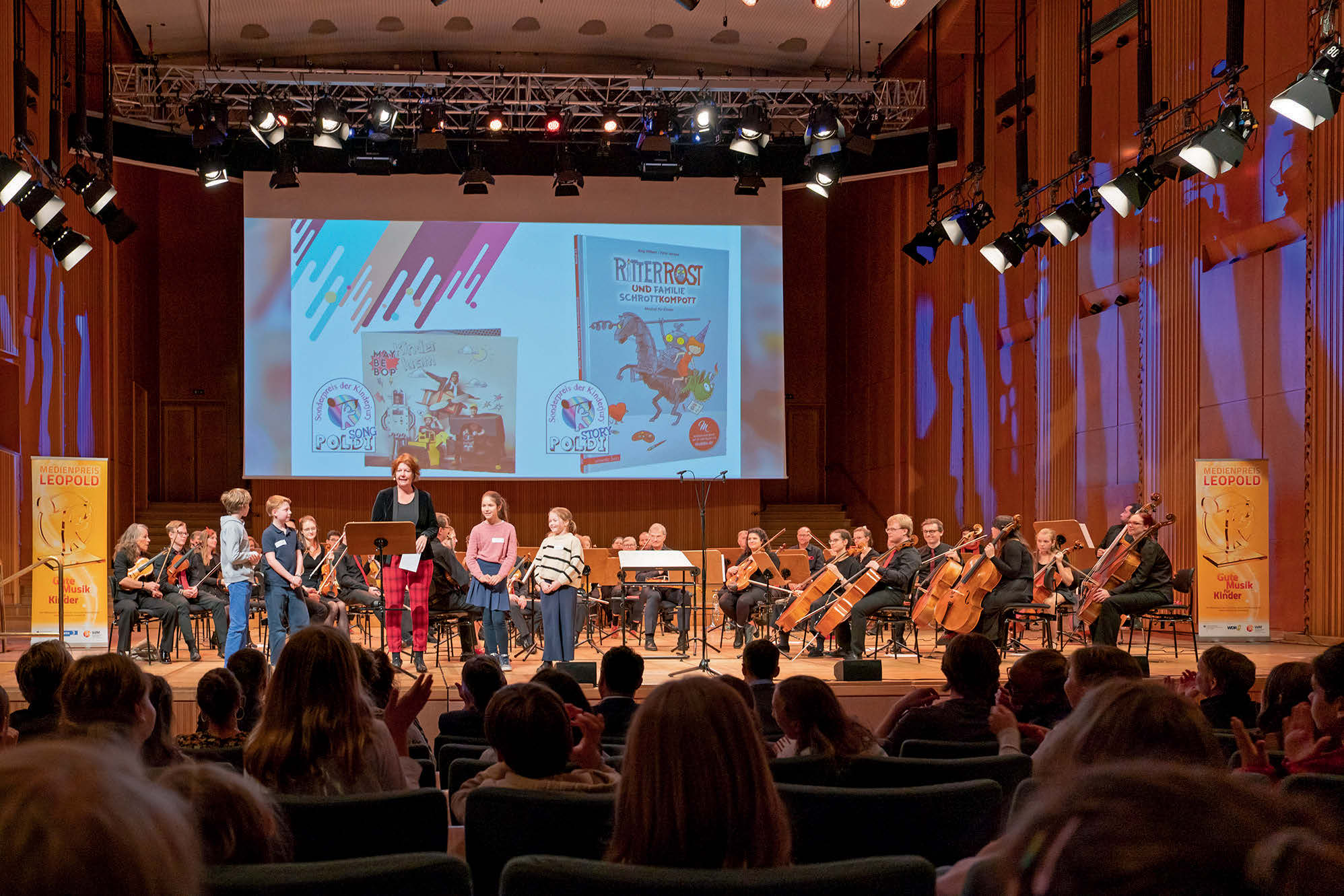 Eine Bühne mit Kinderbüchern an die Rückwand projiziert. Vorne stehen ein paar Kinder dahienter ein Orchester.