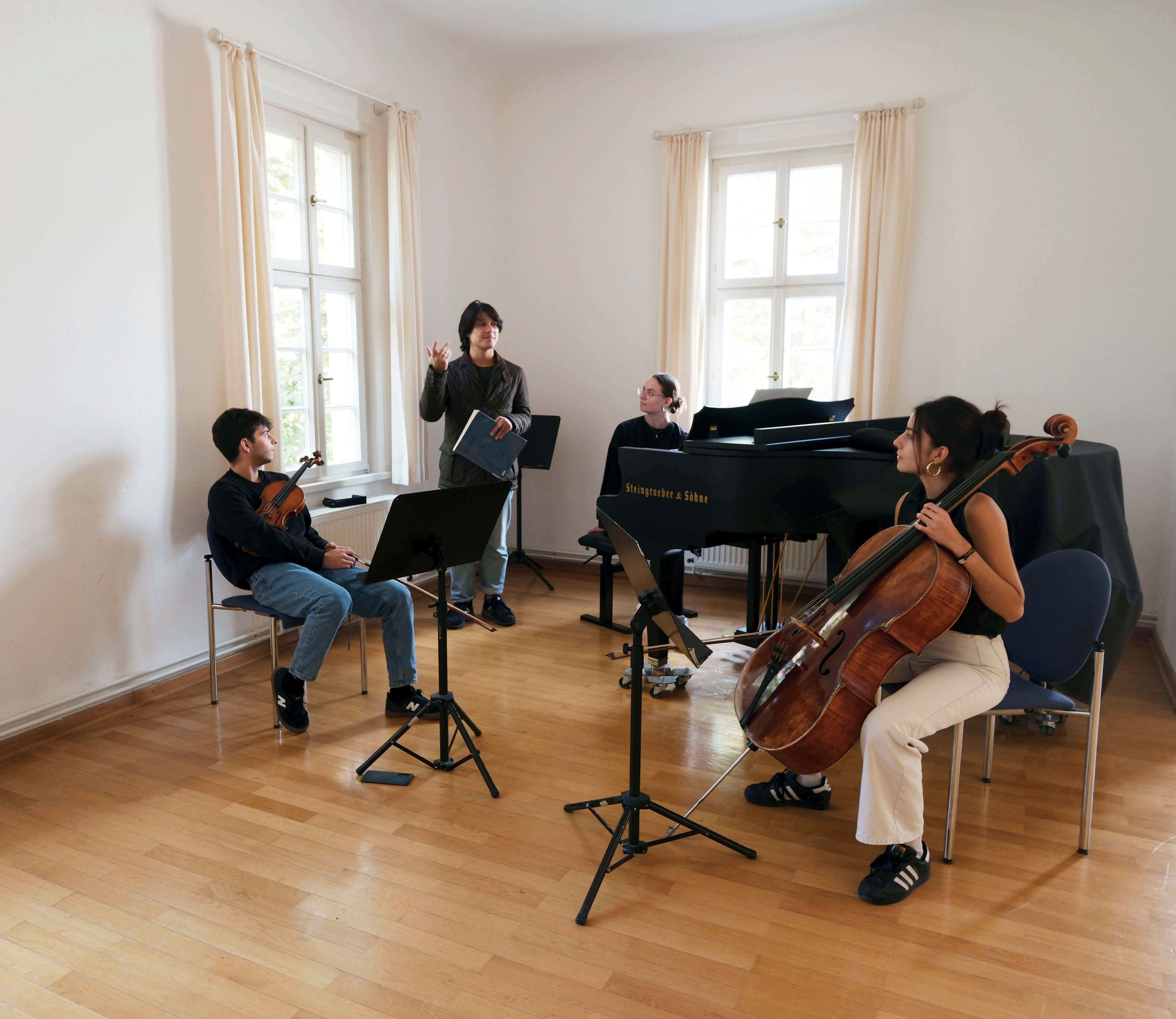 Ein Klaviertrio in einem kleinen ebenfalls weiß verputzten Raum mit creme-farbenen Vorhängne und glänzemdem Parkett. Allerdings kleiner und ohne sichtbare Deckemalerei. Der Dozent steht in der Ecke des Raumes, quasi hinter dem Trio und spricht von dort aus zu ihnen.