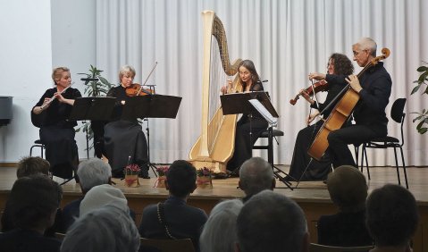 Auf einer Bühne mit weißem Vorhang sieht man von links nach rechts: Die Flötistin, die Violinistin, die Harfenistin, die Bratschistin und den Cellisten. Alle in Schwarz.