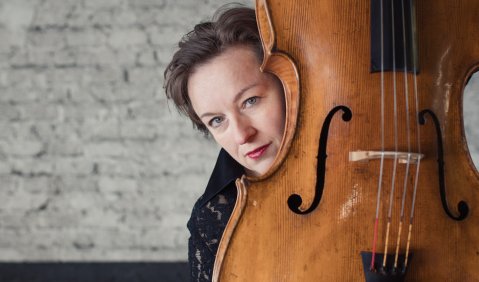 Die Cellistin Joanna Sachryn guckt hinter ihrem Cello hervor. Im Hintergrund eine graue gemauerte Wand.