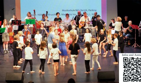 Viele Kinder und Jugendliche sind auf einer Bühne zusammen. Im Halbkreis hinter ihnen steht eine Band oder ein Ensemble, teilweise im Rollstuhl.