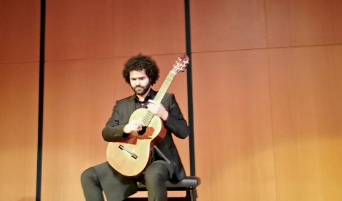 Ein junger Mann mit dichten dunklen Locken und Bart, scharzem Anzug und einer Konzertgitarre. Die sichtbare Wand des Saals hinter ihm ist Holzvertäfelt.