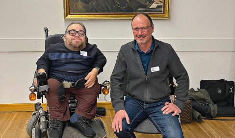 Zwei Männer sitzen in einer Galerie vor einem gemalten Porträt. Der linke, überdurchschnittlich klein, im Rollstuhl, der rechte auf einem Stuhl ohne Rollen.
