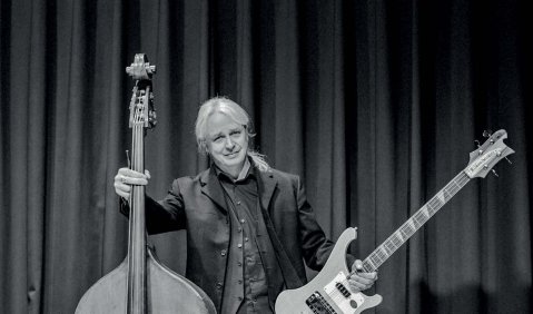 Das Bild ist in Schwarz-Weiß. Es zeigt einen Mann mittleren Alters mit hellen Haaren der mit einer Hand einen Kontrabass hält mit der anderen einen E-Bass. Dahinter ein Bühnenvorhang.