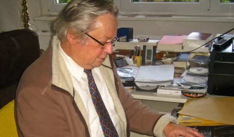 Ein Mann höheren mittleren Alters in brauner Jacke und Hemd und Krawatte an einem E-Piano in einem Arbeitszimmer.