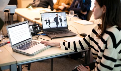 Ein Studentin sitzt hinter zwei Laptops. Einer zeigt Text, der andere ein Tanzvideo. Im Hintergrund sitzen an anderen Tischen noch andere Studierende.