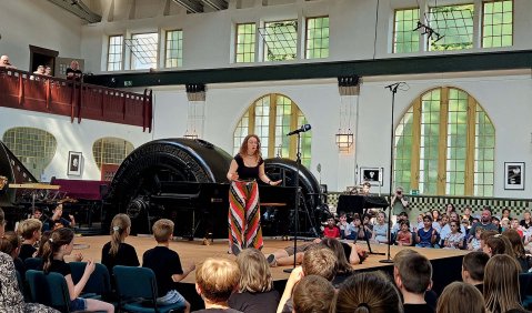 In einer Halle, eines alten Kraftwerks (weißgestrichene Wände, hohe rundbogen Fenster mit Säulen davor). In der Mitte ist eine Bühne umringt von Kindern mit der Schauspielerin in buntem Rock auf der Bühne.