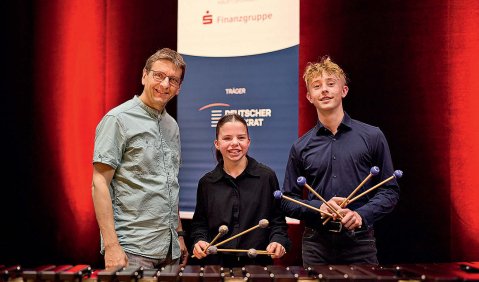 Drei Personen vor einem Marimba. In der mitte die kleinere Antonia Körber in schwarzem Hemd und Mallets in beiden Händen. Rechts von ihr in blauem Hemd und ebenfalls mit Mallets Florenz Richter und links ihr Lehrer, Gregor Daszko. Im Hintergrund ein Jugend musiziert Aufsteller.