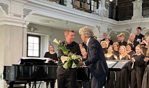 In einem altertümlichen weißen Festsaal auf einer etwas erhöhten Bühne werden dem genannten Blumen überreicht. Im Hintergrund steht ein Chor. Die beiden Herren, die die Blumen austauschen sind schwarz gekleidet mit grauen Haaren.