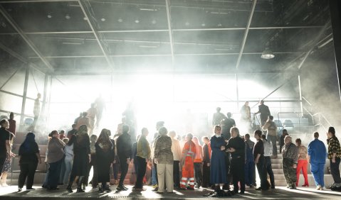 Das Bühnenbild zeigt eine bunte Menschenmenge, die in einer Halle mit Blick ins hell gleißend leuchtend Freie steht.