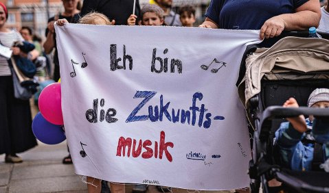 Zwei nicht ganz auf dem Foto gezeigte Menschen, darunter ein verdecktes Kind, tragen ein beschriftetes Laken: "Ich bin die Zukunftsmusik"