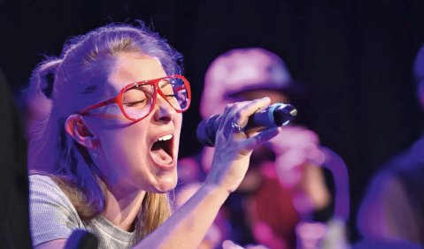Eine junge Frau mit roter Brille singt engagiert im leicht lila-stichigen Rampenlicht. Im Hintergrund sind verschwommen mehrere Background-Sänger:innen zu sehen.
