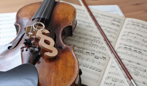 Eine Geige liegt, auf Noten gebettet, auf einem Tisch. Der Bogen daneben, auf der Geige ein hölzernes Paragraphen-Zeichen.