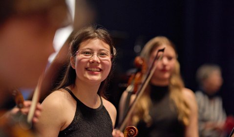 Eine Jugendliche mit langen dunklen Haaren, Brille, einem Schwarzen Kleid, einer Bratsche und einem Lächeln. Im Hintergrund und unscharf im Vordergrund zwei Ensemble Kolleg:innen.