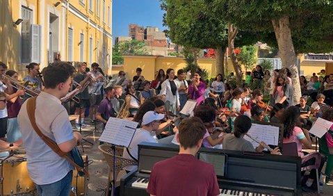 Ein lockerer Open-Air-Auftritt eines großen buntbesetzten Ensembles. Die Sonne scheint, die "Bühne" ist der beschattete Platz vor einem herrschaftlich-gelben Altbau.