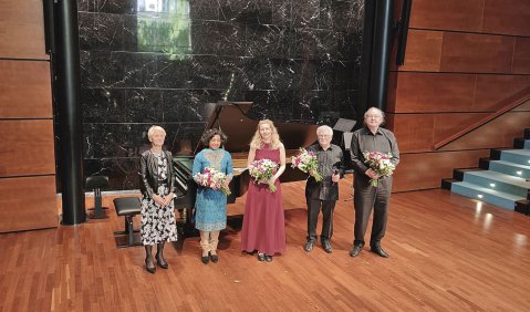 Die beteiligten stehen in farbiger, nicht knalliger, Konzertkleidung mit Blumen in der Hand vor dem Flügel in dem holvertäfelten Saal.