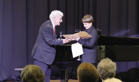 Ein älterer Herr mit weißem Haar und Anzug übergibt einem Jungen (ebenfalls mim Anzug) eine Urkunde. Im Hintergrund ein Flügel und Vorhänge.