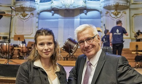 Die beiden, die jüngere Frau in locker-schicker Kleidung, und der ältere Mann in Anzug mit Krawatte stehen nebeneinander im Hintergrund eine Orgel.