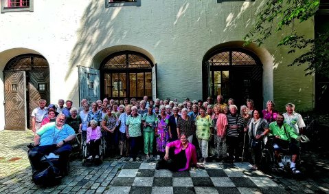 Ein Chor in bunten Klamotten in einem Innenhof. Einige Chormitglieder sitzen in Rollstühlen.