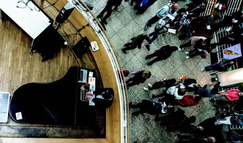 Blick aus der Vogelperspektive: Es wirkt wie ein Foyer, mit einer Art Orchestergraben in dem ein Flügel und vereinzelte Musiker*innen stehen. Auf der höheren Foyerebene isnd diverse Menschen, die verstreut herumstehen.