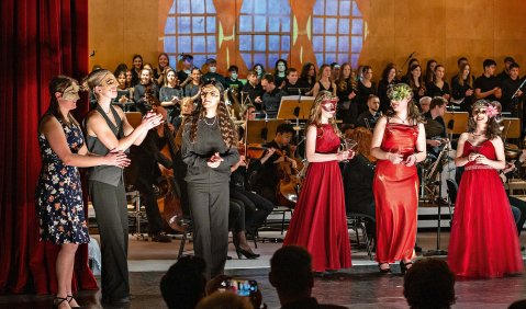 In einem Saal eine halbszenische Aufführung. Vorne auf der Bühne stehen 6 Sängerinnen, drei dunkel und drei Rotbekleidet mit venizianischen Masken im Gesicht. Im Hintergrund Orchester und Chor.