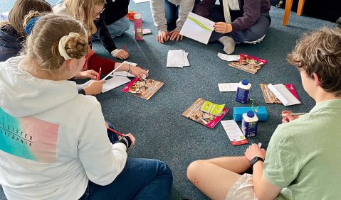 Eine Gruppe Jugendlicher sitzen auf dem Boden im Kreis und arbeiten zusammen mit Zetteln und anderen Materialien.