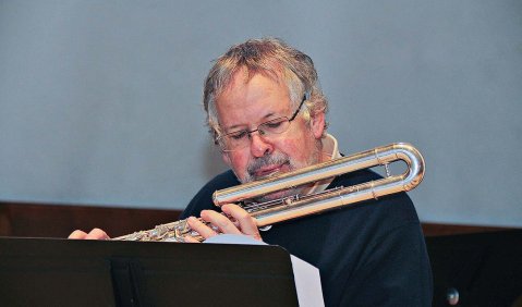 Ein Mann mit leicht schütterem grauen Haar an einer Bass-Querflöte