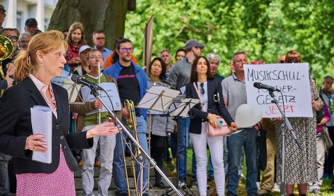 Eine Frau mittleren Alters steht unter freiem Himmel an einem Mikrofon. Im Hintergrund sieht man einige Menschen, manche mit Instrument, andere mit Protestschildern zusammenstehen.