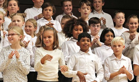 Ein Kinderchor mit Kindern verschiedenster Hautfarben und weißen Oberteilen und schwarzen Hosen.