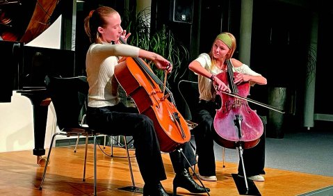 Zwei junge Cellistinnen auf einer kleinen Holzbühne in einem Foyer. Hinter ihnen ein Flügel.