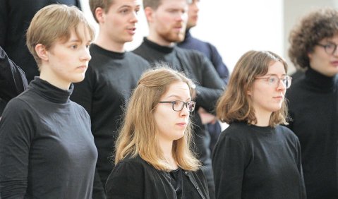 7 Personen aus einem größeren Chor. Sie schauen etwa im 45°-Winkel von der Kamera weg. In schwarzer Kleidung.