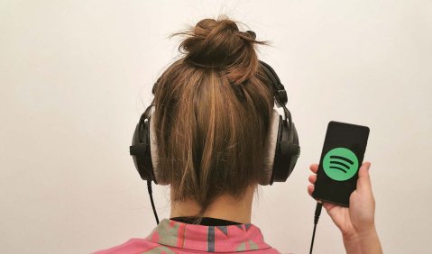 Ein Mensch mit zum Dutt hochgebundenen Haaren und Kopfhörer steht mit dem Rücken zu Kamera. Über die Schulter kann man auf das Smartphone gucken, an dass der Kopfhörer angeschlossen ist. Es zeigt das Spotify-Logo aus grünem Kreis mit drei abwärtsgebogenen Strichen.