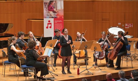 Eine holzvertäfelte Bühne darauf 8 jugendliche Cellist*innen und in ihrer Mitte eine Sängerin.