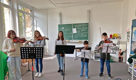 5 Kinder unterschiedlichen Alters, Geschlechts und Haarfarben spielen Geige in einem Klassenzimmer.