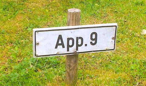 Nahaufnahme eines kleinen Schildes auf einer Wiese, um einen Parkplatz zu benennen. Aufschrift: App. 9.