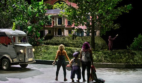 Drei Menschen stehen vor dem Garten vor dem Bayreuther Festspielhaus auf der Straße. Links ein alter Kastenwagen-Bus.